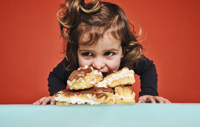 Closeup-Porträt eines fröhlichen kleinen Mädchens, das süße Eclairs mit Schokolade genießt und dabei wegschaut, während es auf dem Tisch vor einem roten Hintergrund sitzt - ADSF09489