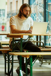 Junger, bärtiger, gut aussehender Mann, der in einem Café sitzt und arbeitet und ein Notebook auf dem Tisch hält - ADSF09494