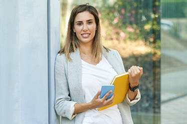 Businesswoman with notebook and smart phone leaning on wall - KIJF03194