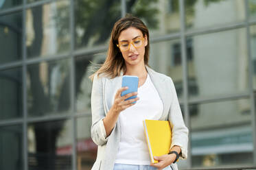 Businesswoman text messaging through smart phone outside office building - KIJF03197