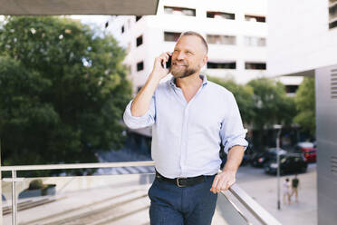 Lächelnder Geschäftsmann, der auf dem Balkon eines Büros steht und mit seinem Handy telefoniert - DGOF01194