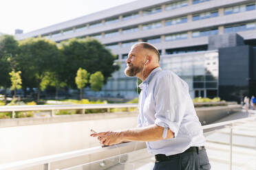 Businessman looking up while listening music through mobile phone at balcony - DGOF01199