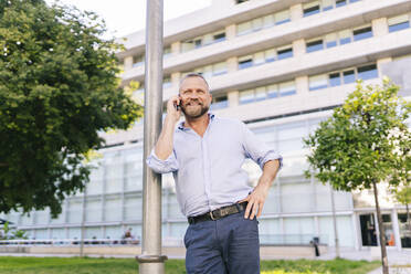 Lächelnder Geschäftsmann, der mit seinem Handy telefoniert, während er sich auf einen Pfahl im Büropark stützt - DGOF01203