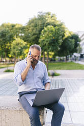 Geschäftsmann, der mit einem Smartphone spricht, während er einen Laptop im Büropark benutzt - DGOF01208