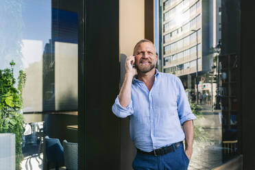 Smiling businessman talking on mobile phone while leaning on wall - DGOF01213