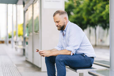 Geschäftsmann benutzt sein Smartphone, während er auf einer Bank am Bahnhof sitzt - DGOF01230