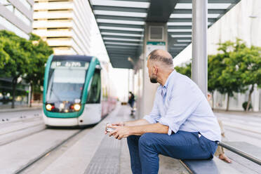 Geschäftsmann schaut auf die Straßenbahn, während er auf einer Bank am Bahnhof in der Stadt sitzt - DGOF01231