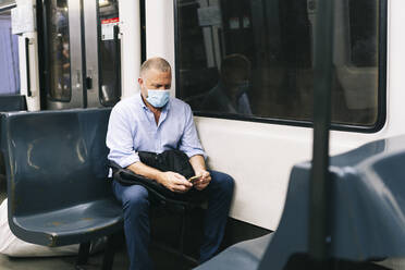 Geschäftsmann mit Gesichtsmaske, der während der Fahrt im Zug sein Smartphone benutzt - DGOF01235