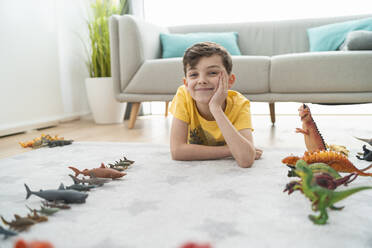 Lächelnder Junge mit Hand am Kinn, der neben Spielzeugtieren auf dem Teppich im Wohnzimmer liegt - SNF00450