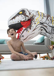 Hemdloser fröhlicher Junge, der mit einem großen Spielzeugdinosaurier spielt, während er im Wohnzimmer sitzt - SNF00458