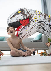 Shirtless cheerful boy playing with large toy dinosaur while sitting in living room - SNF00458