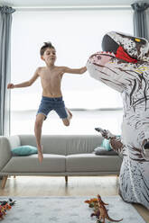 Shirtless boy jumping from sofa while fighting with large toy dinosaur - SNF00459