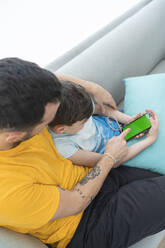 Father with son using smart phone while relaxing on sofa at home - SNF00475