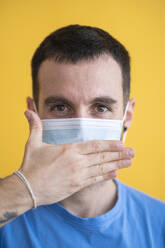 Close-up of mid adult man wearing mask covering mouth with hand against yellow background - SNF00481