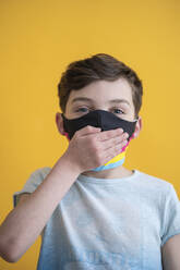 Nahaufnahme eines Jungen mit Maske, der den Mund mit der Hand bedeckt, vor gelbem Hintergrund - SNF00484
