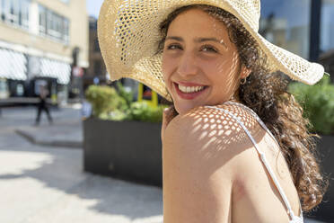 Nahaufnahme einer lächelnden schönen Frau mit Hut, die an einem sonnigen Tag in der Stadt sitzt - WPEF03219
