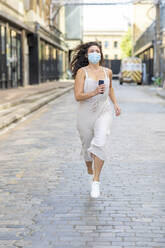 Young woman wearing mask running on street in city - WPEF03224