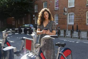 Schöne Frau, die über ihr Smartphone an einer Parkstation ein Fahrrad mietet - WPEF03231