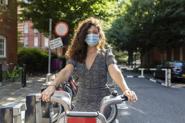 Junge Frau mit Maske sitzt auf einem Fahrrad in einer Parkstation in der Stadt - WPEF03234