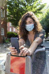 Young woman wearing mask taking selfie with smart phone while sitting on bicycle at parking station - WPEF03235