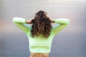 Junge Frau in grünem Crop-Top mit Händen in den Haaren posiert gegen eine graue Wand - WPEF03244