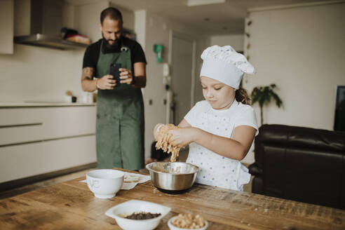 Vater und Tochter backen zu Hause Plätzchen, der Vater macht ein Foto mit dem Smartphone - GMLF00428