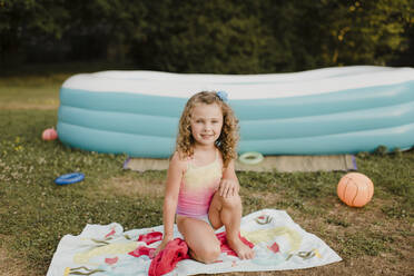 Porträt eines lächelnden Mädchens auf einem Handtuch am aufblasbaren Swimmingpool im Garten - SMSF00111