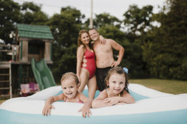 Portrait of two happy girls in an inflatable swimming pool with parents in background - SMSF00129