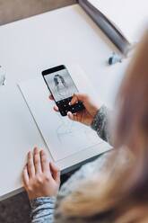 From above crop blond girl taking photo on mobile phone of first sketch on paper at table in office - ADSF09549