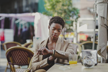 African American elegant woman looking at watch and sitting at table with mobile phone and glass of juice in street cafe on blurred background - ADSF09550