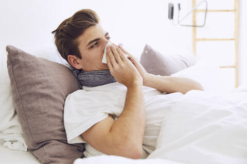 Kranker junger Mann schnäuzt sich die Nase, während er zu Hause im Bett liegt, lizenzfreies Stockfoto