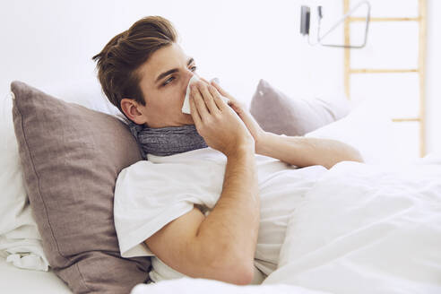 Kranker junger Mann schnäuzt sich die Nase, während er zu Hause im Bett liegt - UKOF00019