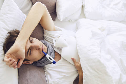 Sick young man with thermometer in mouth resting on bed at home stock photo