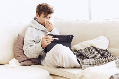 Junger Mann schnäuzt sich die Nase, während er ein digitales Tablet auf dem Sofa zu Hause benutzt, lizenzfreies Stockfoto