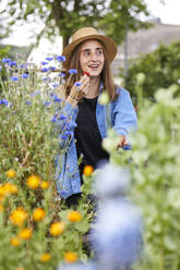 Lächelnde Frau mit Hut isst Erdbeere, während sie inmitten von Pflanzen im Garten sitzt - UKOF00056