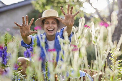 Cheerful woman wearing hat showing dirty hands while sitting amidst plants in garden - UKOF00068