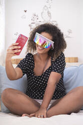 Frau mit mehrfarbiger Sonnenbrille nimmt Selfie mit Smartphone auf Bett zu Hause - SNF00494