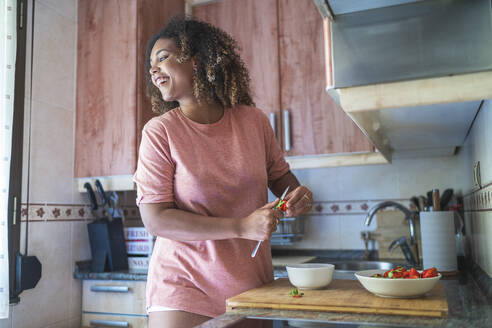 Fröhliche junge Frau, die wegschaut, während sie Erdbeeren auf dem Küchentisch zu Hause schneidet - SNF00496
