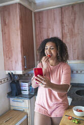 Cheerful young woman eating strawberry while video calling over smart phone in kitchen - SNF00498