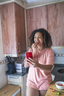 Fröhliche junge Frau, die eine Erdbeere isst, während sie in der Küche einen Videogespräch über ein Smartphone führt - SNF00498