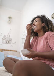 Cheerful young woman eating food while sitting on bed at home - SNF00504