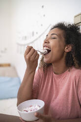 Nahaufnahme einer fröhlichen Frau, die Erdbeeren mit Sahne isst, während sie zu Hause sitzt - SNF00505
