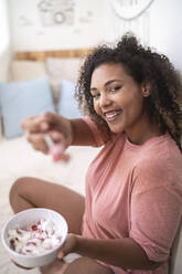 Fröhliche Frau zeigt Erdbeeren mit Sahne, während sie zu Hause auf dem Bett sitzt - SNF00509