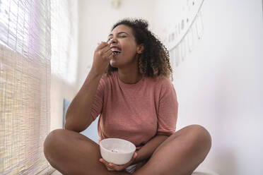 Cheerful woman eating cream and strawberries while sitting by wall at home - SNF00511