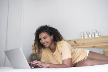Lächelnde junge Frau mit lockigem Haar, die einen Laptop benutzt, während sie zu Hause auf dem Bett liegt - SNF00512