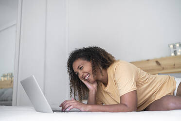 Fröhliche Frau mit lockigem Haar, die einen Laptop benutzt, während sie zu Hause auf dem Bett liegt - SNF00513