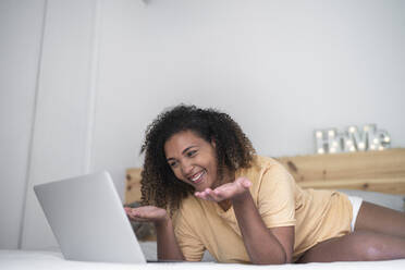 Fröhliche junge Frau bei einer Videokonferenz über einen Laptop, während sie zu Hause auf dem Bett liegt - SNF00514