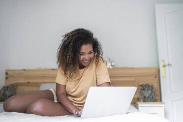 Lächelnde junge Frau mit lockigem Haar, die einen Laptop benutzt, während sie zu Hause auf dem Bett sitzt - SNF00515