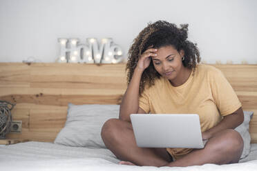 Junge Frau mit lockigem Haar, die auf dem Bett sitzend einen Laptop benutzt - SNF00520