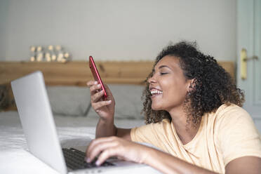 Fröhliche junge Frau mit Smartphone und Laptop auf dem Bett zu Hause - SNF00527